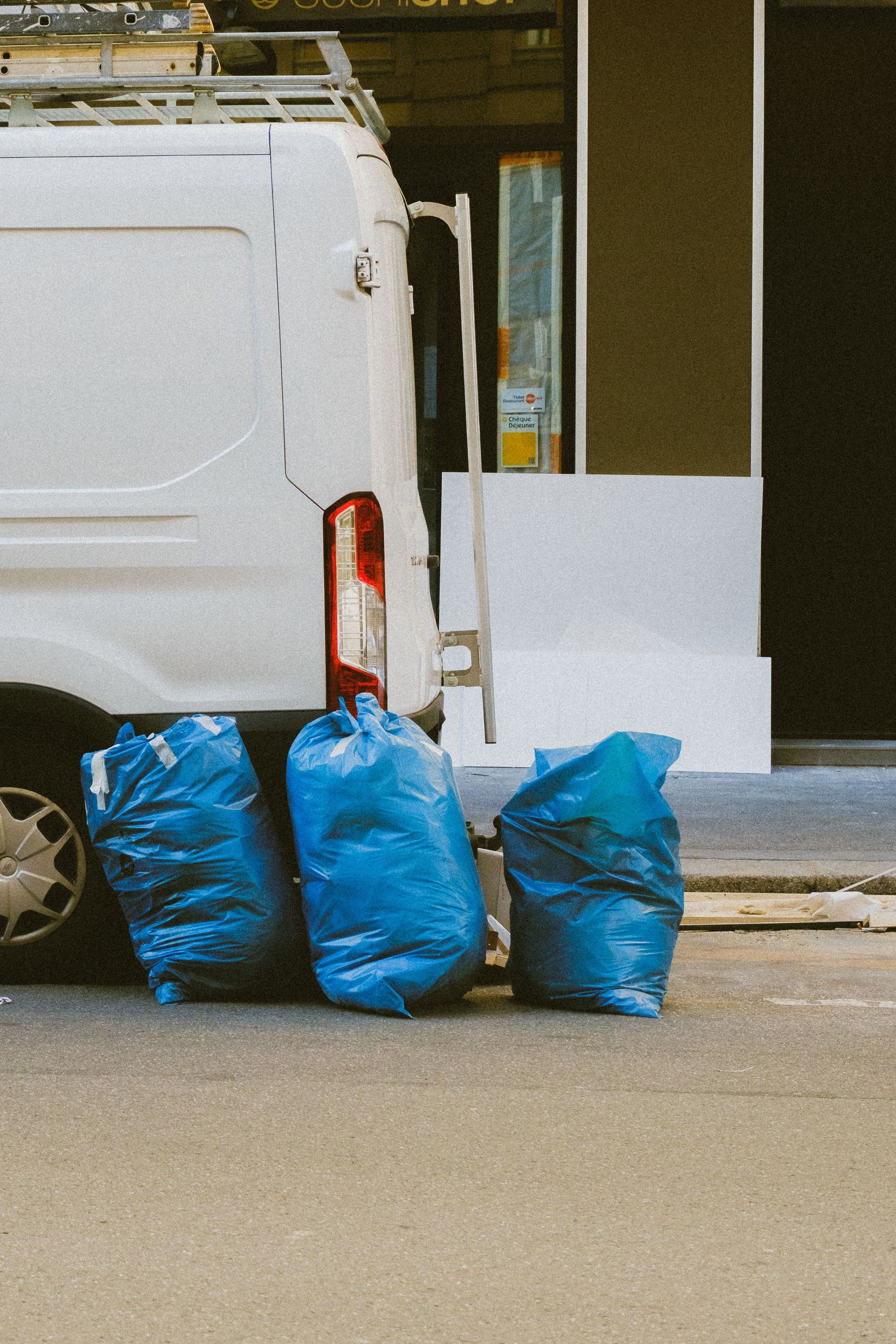 Three blue garbage bags next to a white van in an urban setting. Daytime photo.